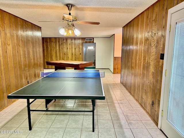 rec room with a textured ceiling, wooden walls, ceiling fan, and light tile patterned floors