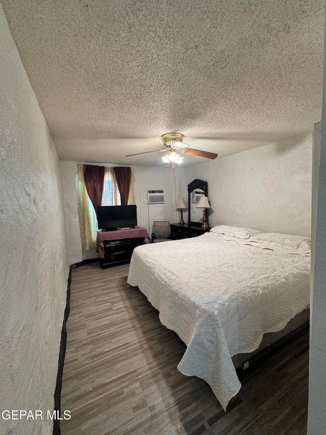 bedroom with a wall unit AC, a textured ceiling, hardwood / wood-style flooring, and ceiling fan