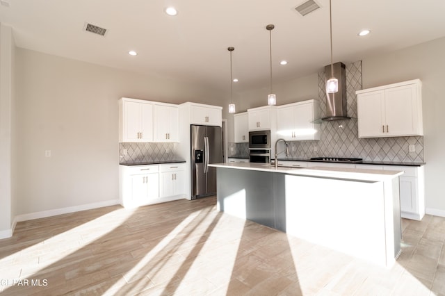 kitchen featuring stainless steel appliances, wall chimney range hood, pendant lighting, white cabinets, and an island with sink