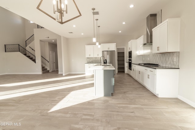 kitchen featuring stainless steel appliances, wall chimney range hood, backsplash, pendant lighting, and a center island with sink