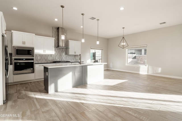 kitchen featuring hanging light fixtures, stainless steel appliances, wall chimney range hood, an island with sink, and white cabinets