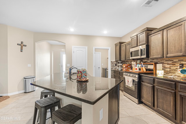 kitchen featuring a kitchen bar, appliances with stainless steel finishes, decorative backsplash, a center island with sink, and dark stone countertops