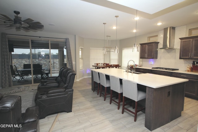 kitchen with sink, wall chimney exhaust hood, an island with sink, stainless steel gas stovetop, and dark brown cabinets