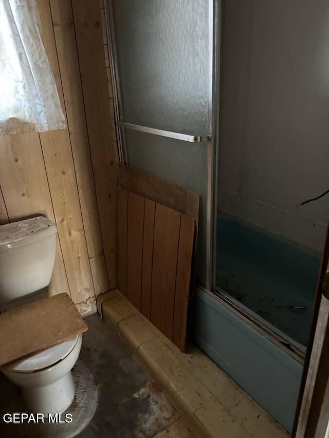 bathroom with shower / bath combination with glass door, toilet, and wood walls