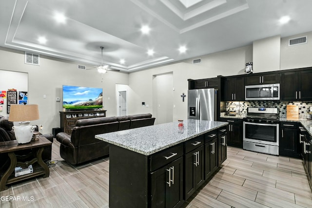 kitchen with stainless steel appliances, light stone counters, a tray ceiling, decorative backsplash, and a kitchen island