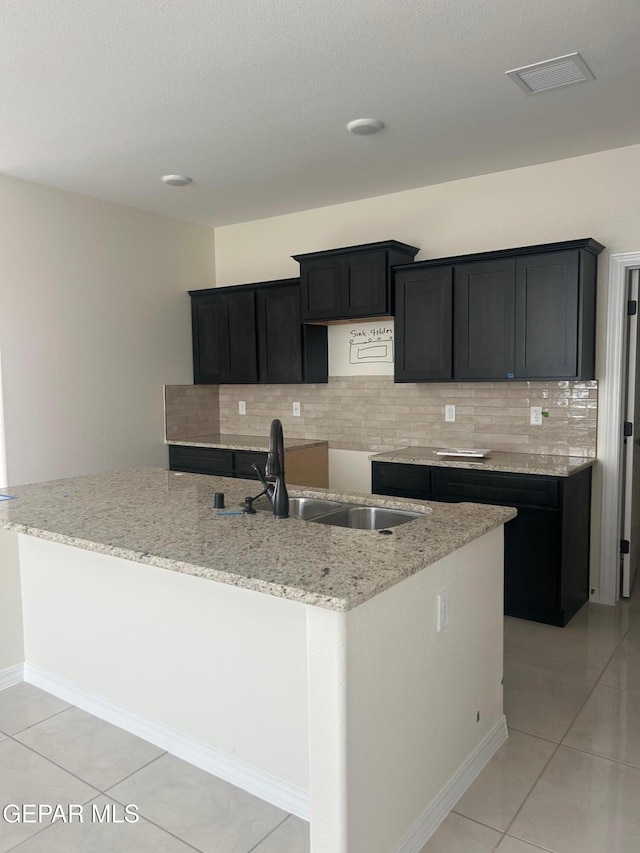 kitchen featuring sink, tasteful backsplash, light tile patterned floors, light stone counters, and a center island with sink