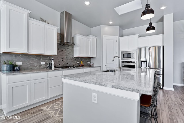 kitchen with white cabinetry, sink, wall chimney exhaust hood, stainless steel appliances, and a kitchen island with sink