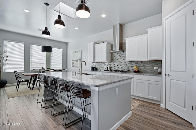 kitchen with a kitchen island with sink, white cabinets, sink, hanging light fixtures, and wall chimney exhaust hood