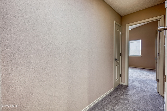 hall with a textured wall, dark carpet, a textured ceiling, and baseboards