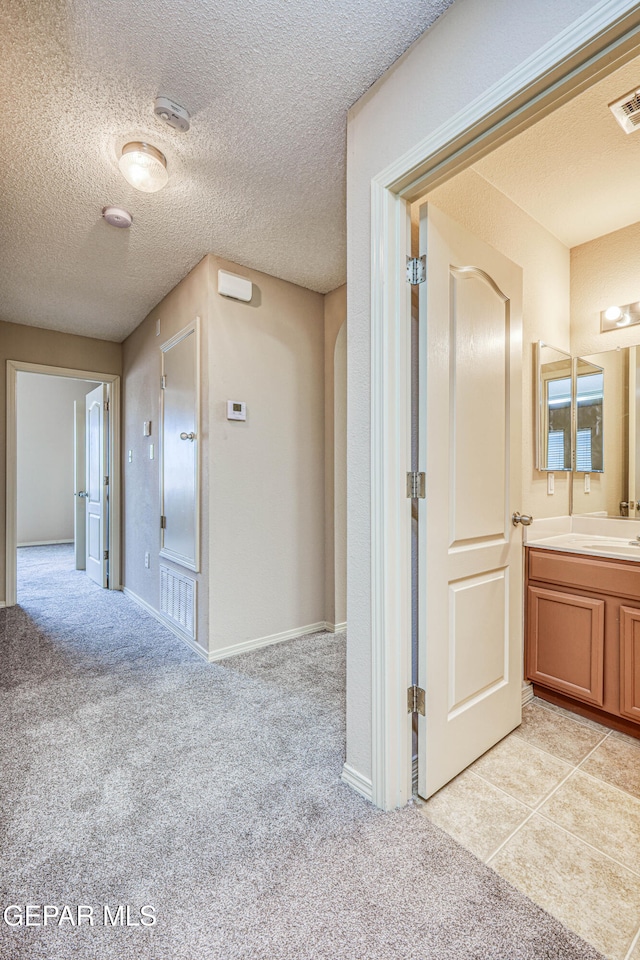 corridor featuring visible vents, light colored carpet, a sink, a textured ceiling, and baseboards