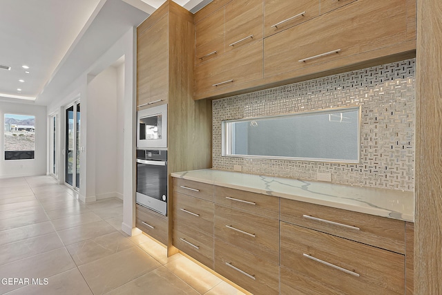 kitchen featuring white microwave, oven, light tile patterned floors, tasteful backsplash, and light stone counters