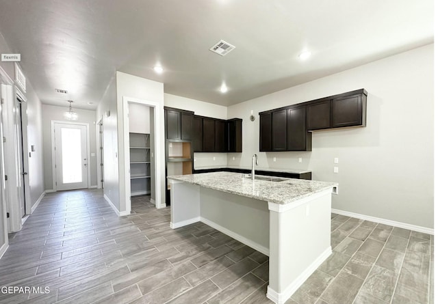 kitchen featuring light stone countertops, sink, dark brown cabinets, a center island with sink, and light wood-type flooring