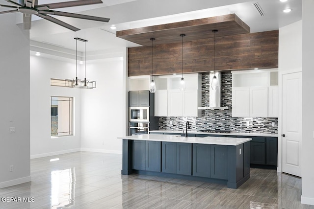 kitchen featuring wall chimney range hood, decorative light fixtures, a center island with sink, white cabinets, and appliances with stainless steel finishes