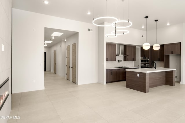 kitchen with pendant lighting, a kitchen island with sink, wall chimney range hood, appliances with stainless steel finishes, and dark brown cabinetry