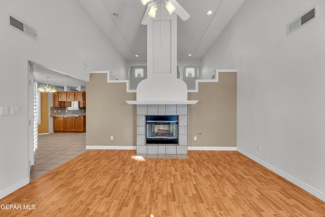 unfurnished living room with ceiling fan with notable chandelier, a tile fireplace, a high ceiling, and light hardwood / wood-style floors