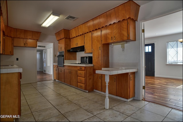 kitchen with tile counters, black oven, stainless steel stovetop, and light hardwood / wood-style flooring