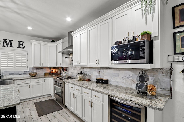 kitchen with light stone countertops, appliances with stainless steel finishes, wall chimney exhaust hood, beverage cooler, and white cabinetry