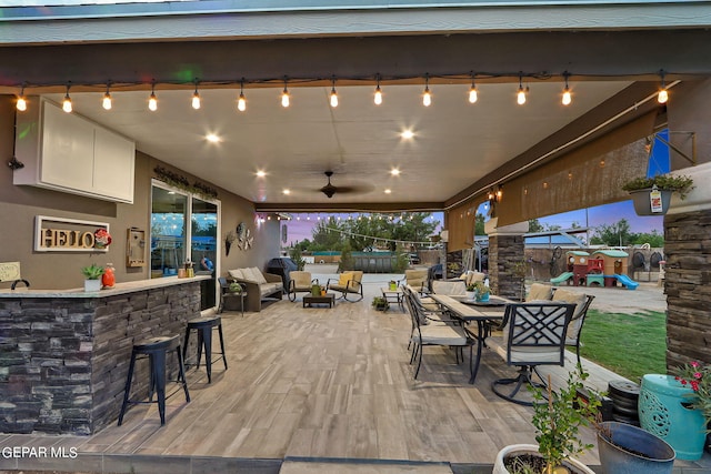 patio terrace at dusk with a bar, an outdoor hangout area, a playground, and ceiling fan