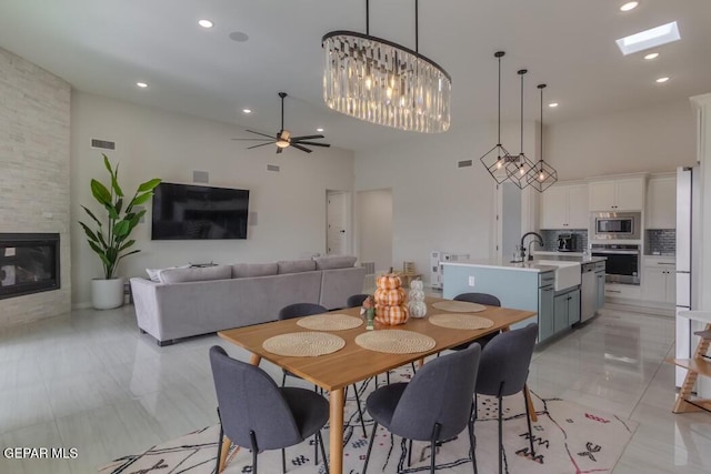 tiled dining space with a towering ceiling, a skylight, ceiling fan with notable chandelier, sink, and a stone fireplace