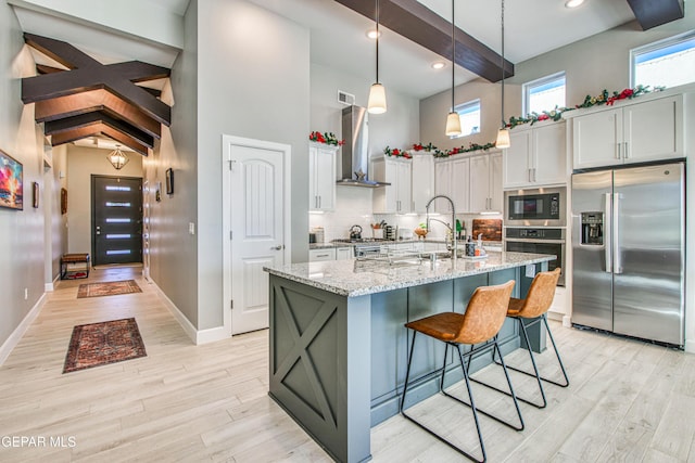 kitchen with beam ceiling, light stone countertops, stainless steel appliances, light hardwood / wood-style flooring, and a center island with sink