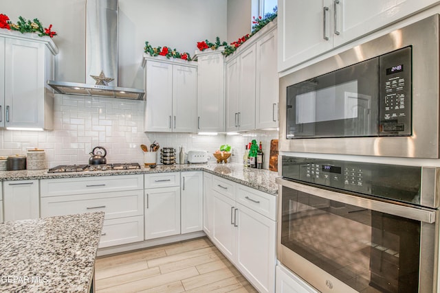 kitchen with white cabinets, stainless steel appliances, light stone counters, and wall chimney exhaust hood