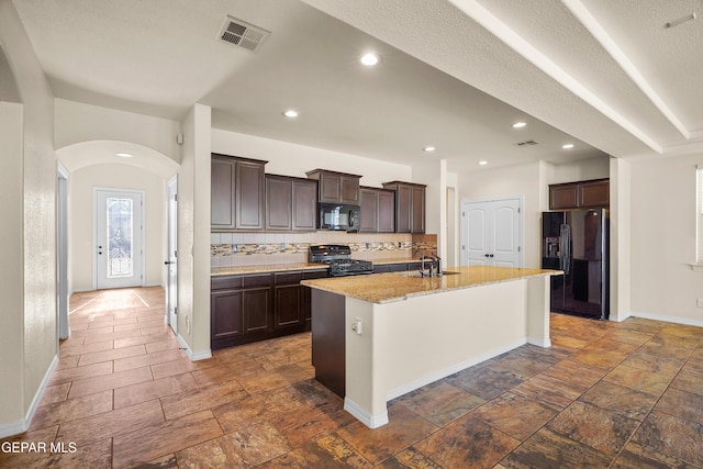 kitchen with dark brown cabinetry, sink, backsplash, a center island with sink, and black appliances