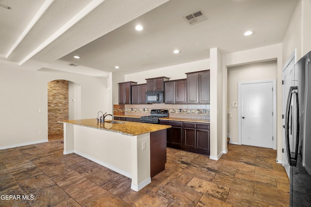 kitchen with decorative backsplash, dark brown cabinets, a kitchen island with sink, sink, and black appliances