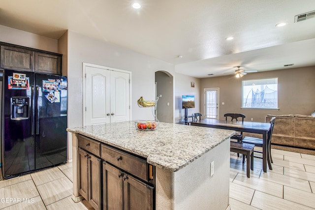 kitchen with light stone countertops, black refrigerator with ice dispenser, dark brown cabinets, ceiling fan, and a center island