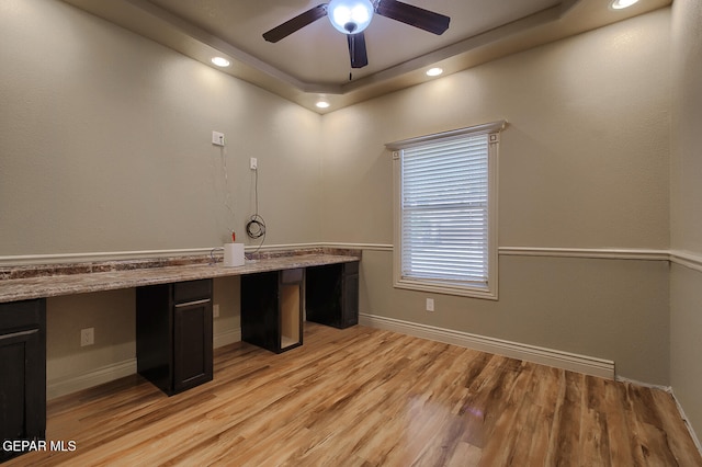 unfurnished office featuring a tray ceiling, ceiling fan, built in desk, and light wood-type flooring