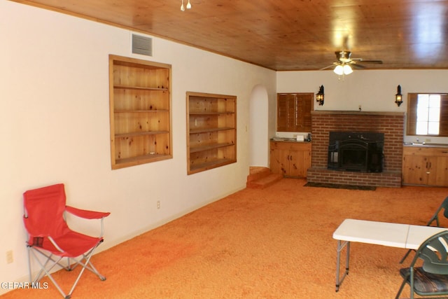 carpeted living room with ceiling fan, crown molding, wooden ceiling, and a brick fireplace