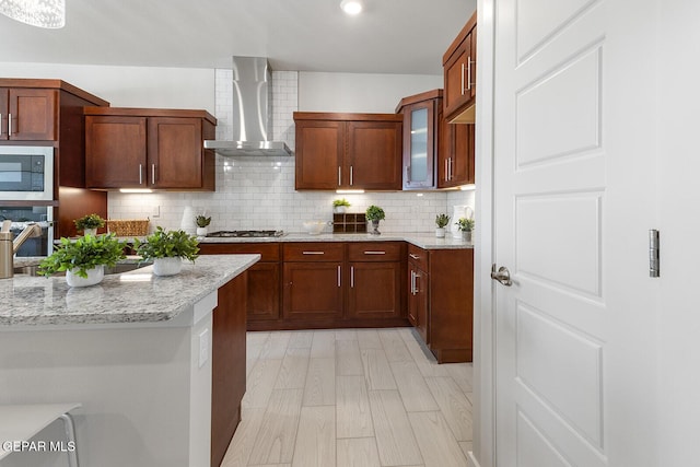 kitchen featuring wall chimney range hood, appliances with stainless steel finishes, tasteful backsplash, light stone countertops, and light wood-type flooring