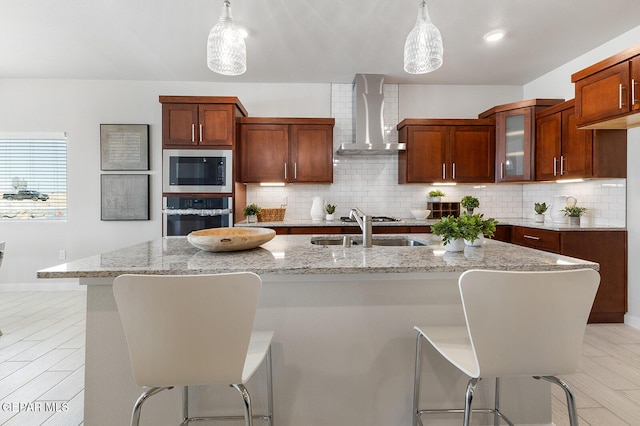 kitchen with pendant lighting, wall chimney range hood, an island with sink, built in microwave, and stainless steel oven