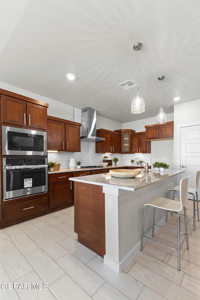 kitchen featuring wall chimney exhaust hood, hanging light fixtures, a center island with sink, stainless steel appliances, and light stone countertops