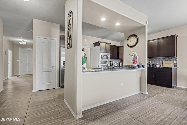 kitchen with dark brown cabinetry, kitchen peninsula, dark stone countertops, decorative backsplash, and appliances with stainless steel finishes