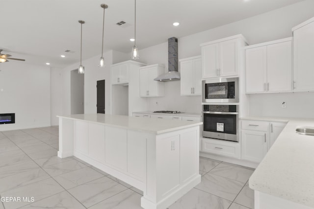 kitchen with wall chimney exhaust hood, decorative light fixtures, a kitchen island, stainless steel appliances, and white cabinets