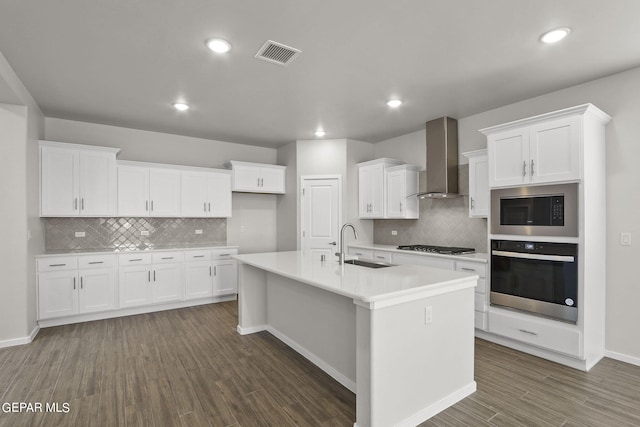 kitchen featuring wall chimney exhaust hood, stainless steel appliances, sink, white cabinets, and an island with sink