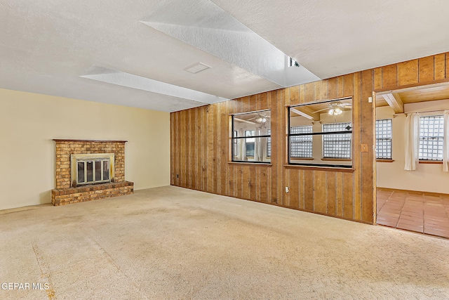 unfurnished living room featuring ceiling fan, a brick fireplace, wood walls, carpet floors, and a textured ceiling