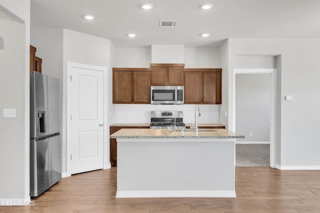 kitchen with light stone counters, sink, stainless steel appliances, and an island with sink