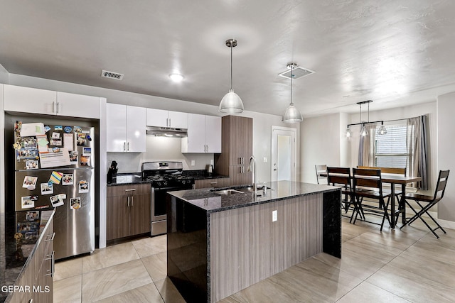 kitchen featuring appliances with stainless steel finishes, dark stone counters, a kitchen island with sink, pendant lighting, and white cabinetry