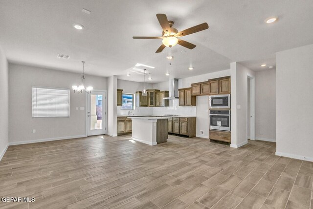 kitchen with a center island, wall chimney exhaust hood, stainless steel appliances, decorative light fixtures, and ceiling fan with notable chandelier