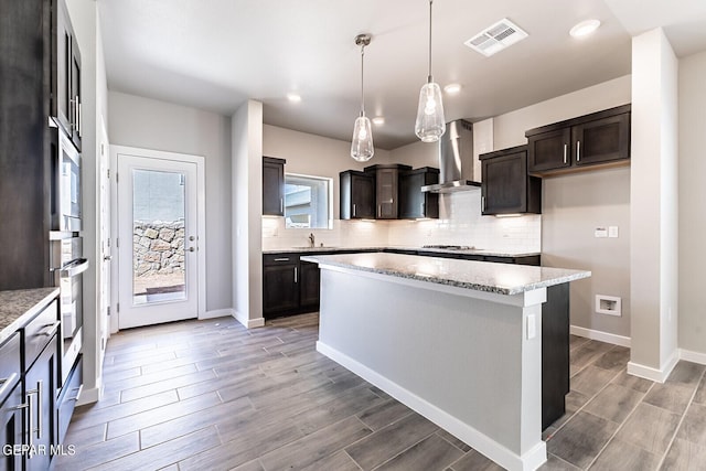 kitchen with wall chimney exhaust hood, stainless steel gas stovetop, dark brown cabinets, a kitchen island, and hanging light fixtures