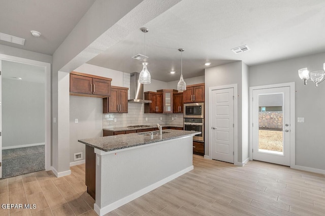 kitchen featuring wall chimney range hood, decorative light fixtures, stone countertops, a kitchen island with sink, and appliances with stainless steel finishes