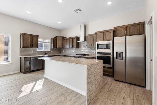 kitchen featuring appliances with stainless steel finishes, light stone counters, sink, wall chimney range hood, and a center island