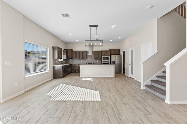 kitchen with wall chimney range hood, hanging light fixtures, a kitchen island, appliances with stainless steel finishes, and light hardwood / wood-style floors