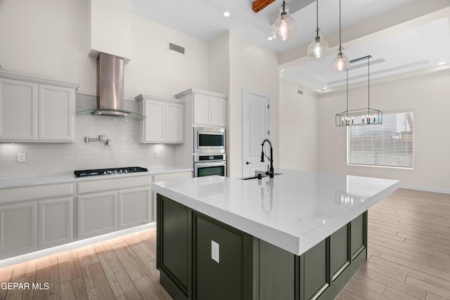 kitchen with white cabinetry, wall chimney exhaust hood, hanging light fixtures, an island with sink, and appliances with stainless steel finishes