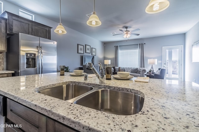 kitchen featuring sink, stainless steel refrigerator with ice dispenser, hanging light fixtures, ceiling fan, and light stone countertops