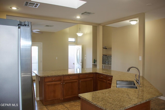 kitchen featuring sink, hanging light fixtures, light stone counters, stainless steel fridge, and a center island with sink