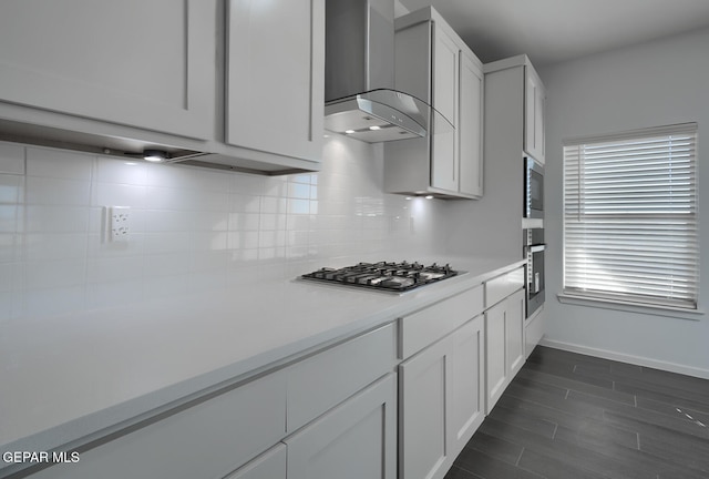 kitchen featuring wall chimney range hood, light countertops, backsplash, baseboards, and appliances with stainless steel finishes