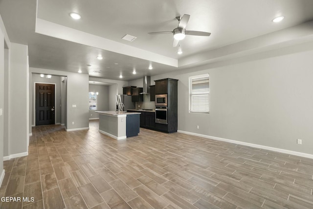 kitchen featuring a raised ceiling, wall chimney exhaust hood, ceiling fan, an island with sink, and appliances with stainless steel finishes