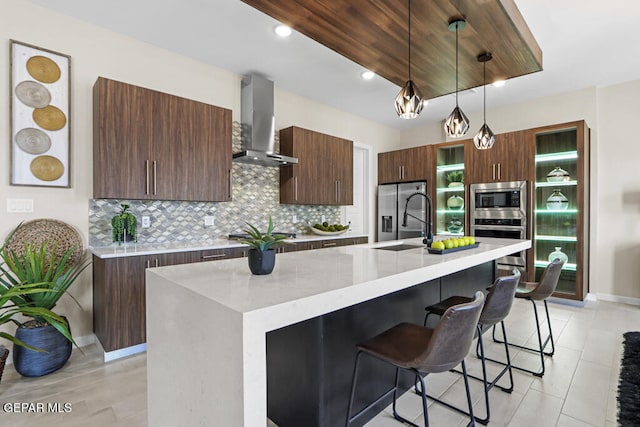 kitchen with stainless steel appliances, hanging light fixtures, wall chimney exhaust hood, and an island with sink